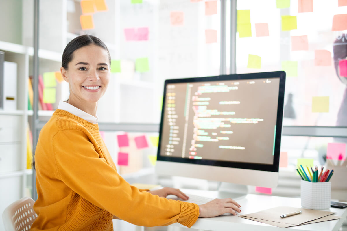 Pretty young webdesigner in casualwear looking at you by her workplace while working over new project