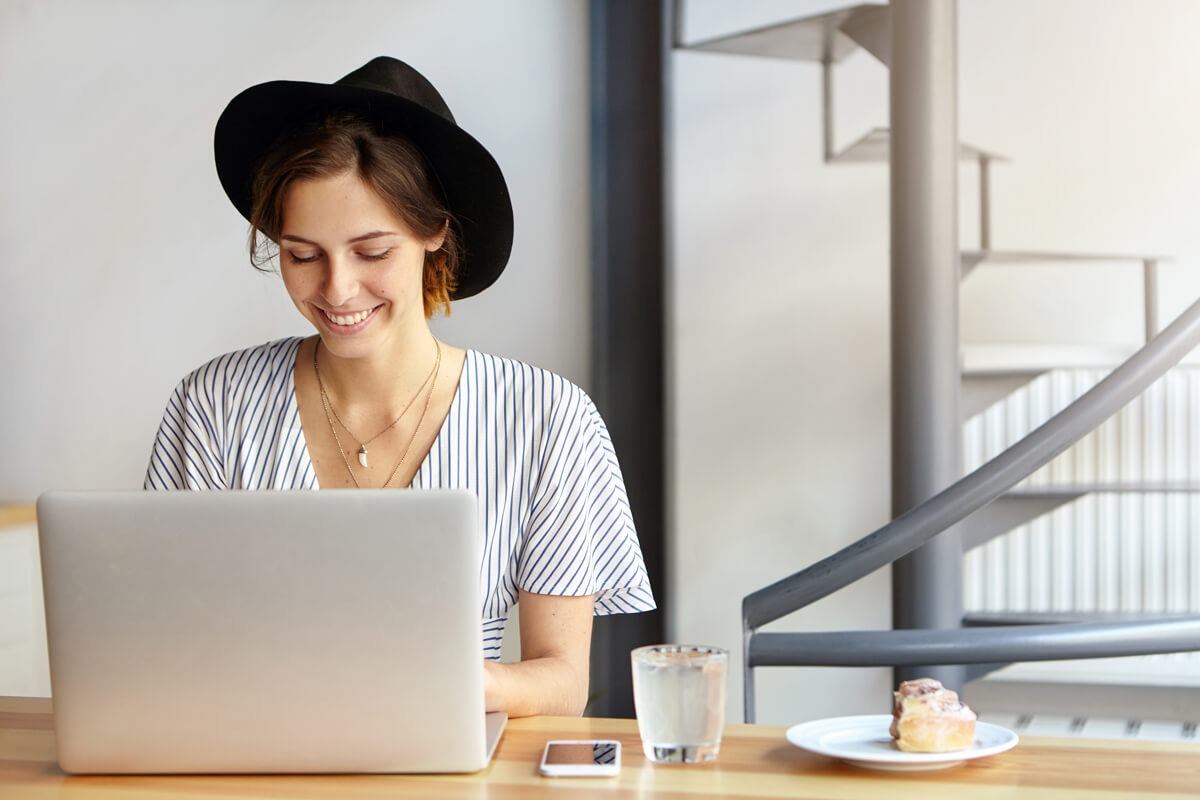 Pretty young female copywriter wearing black hat and stripped blouse working on laptop computer, looking happily typing something sitting at wooden table