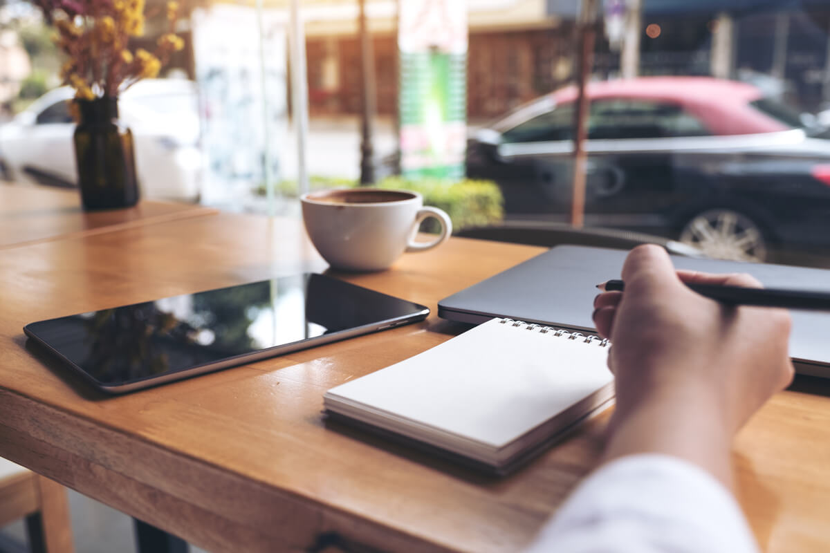 woman's hand writing on a blank notebook with laptop , tablet and coffee cup on wooden table