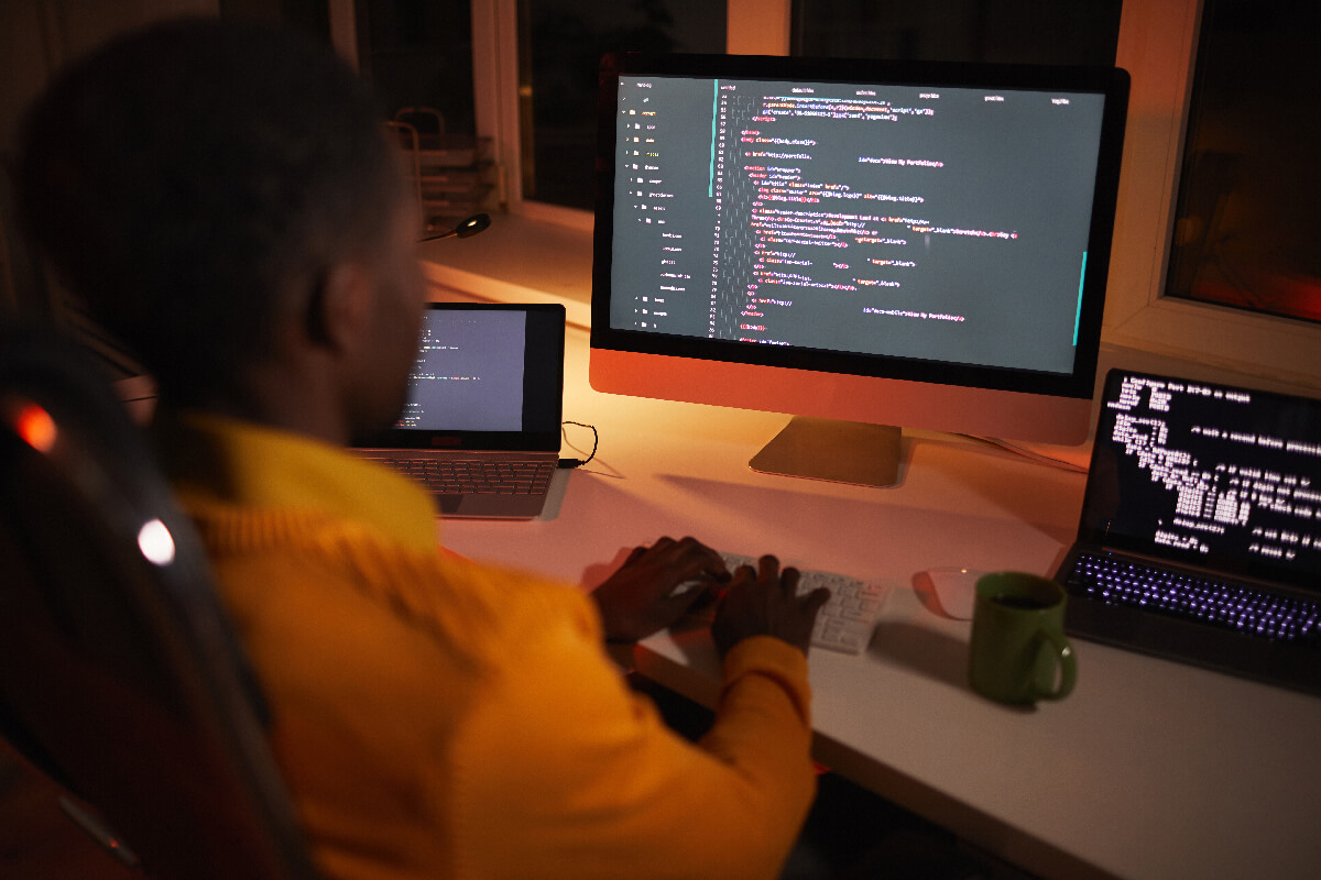 Back view portrait of African-American man writing code while working with multiple computer screens in dark room