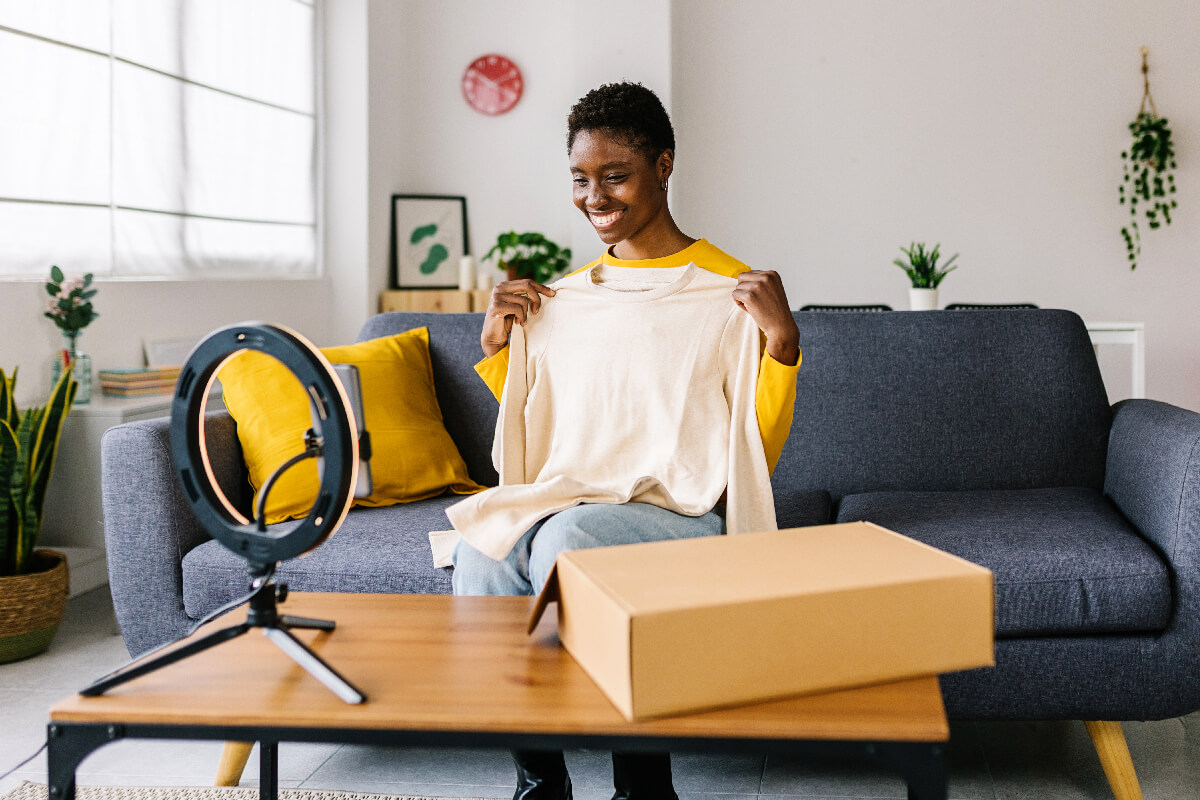 Smiling african american blogger influencer recording video communication with subscribers at home. Young happy woman unpacking parcel and smiling to the smartphone camera