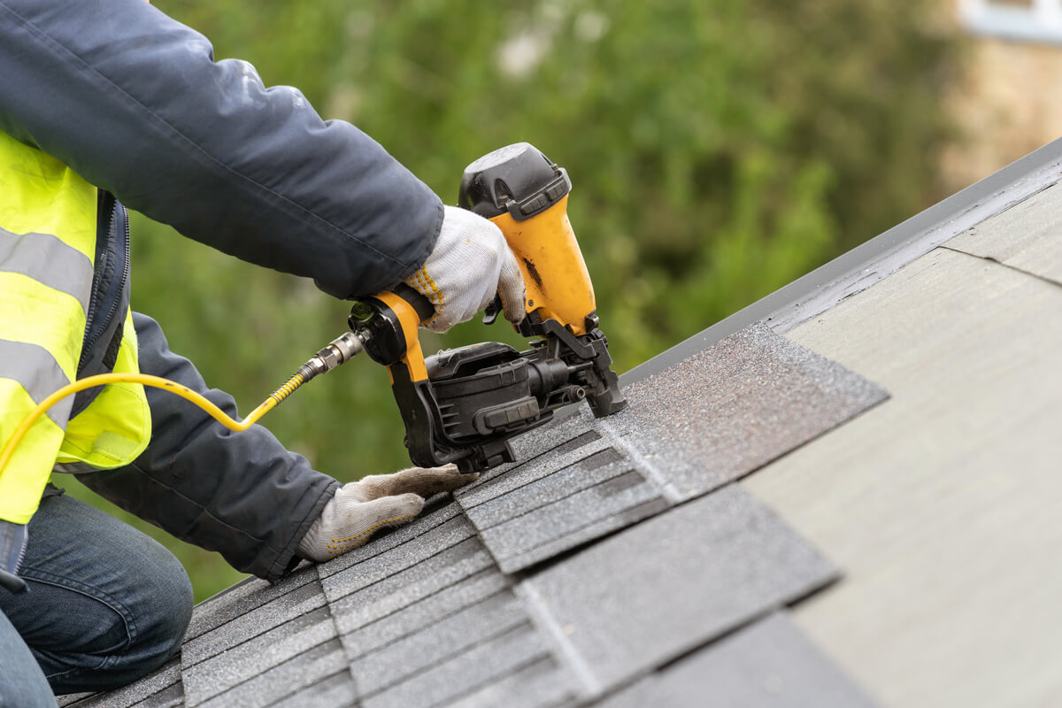 roofer worker in uniform work wear using air or pneumatic nail gun and installing asphalt or bitumen tile on top of the roof under construction house