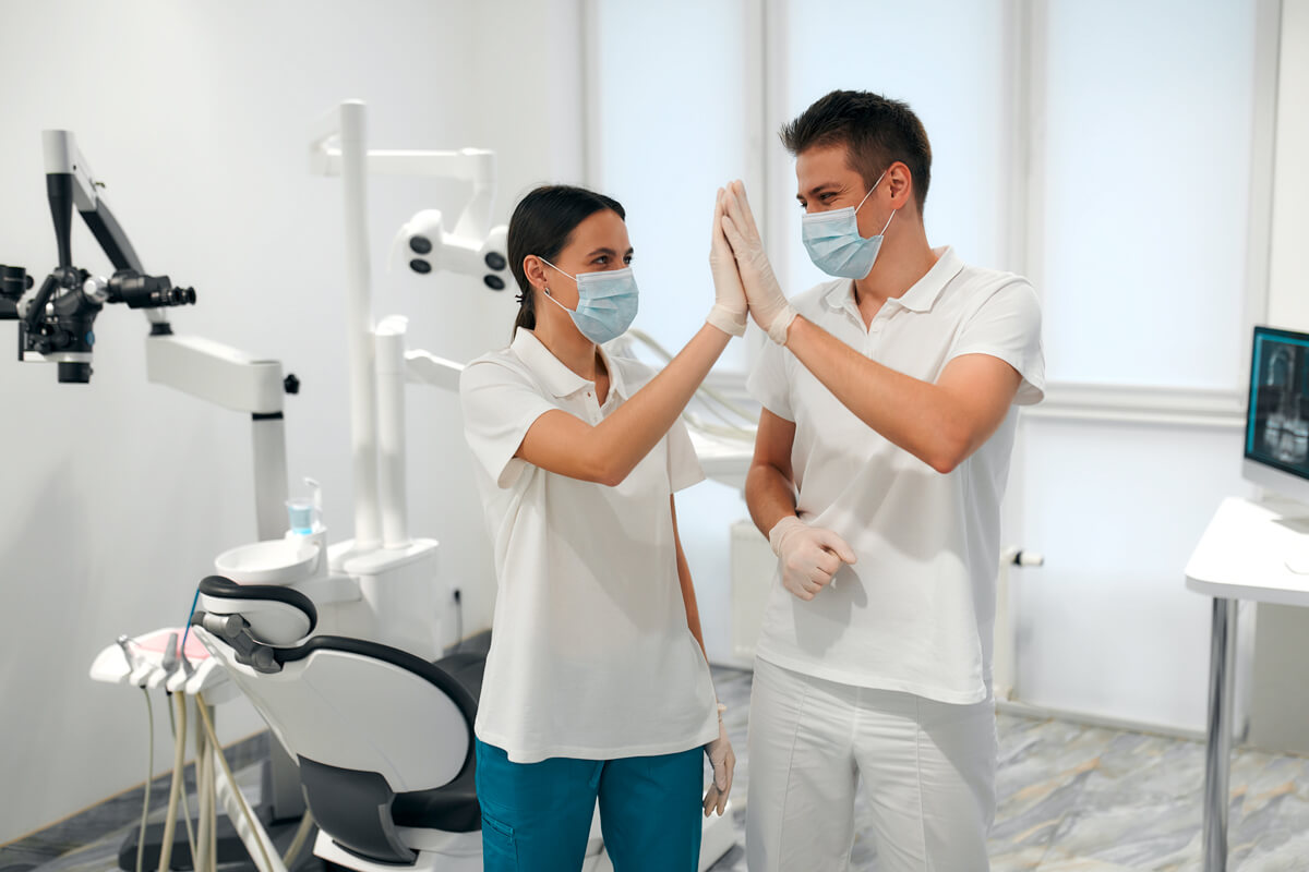 A young dentist and his assistant in a white uniform and medical masks give high five in the dental office. Dentistry and dental care.