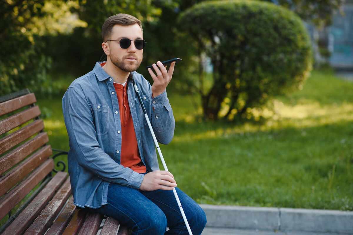 Young blind man with smartphone sitting on bench in park in city.